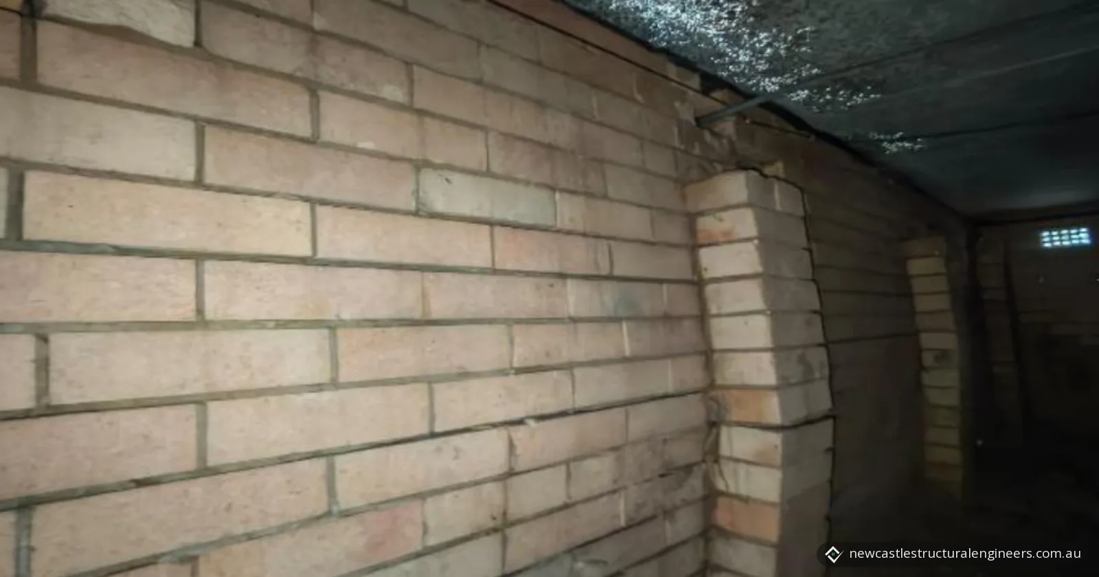 Cracked engaged brick pier bowing away from a brick wall in a dim subfloor, under a concrete slab